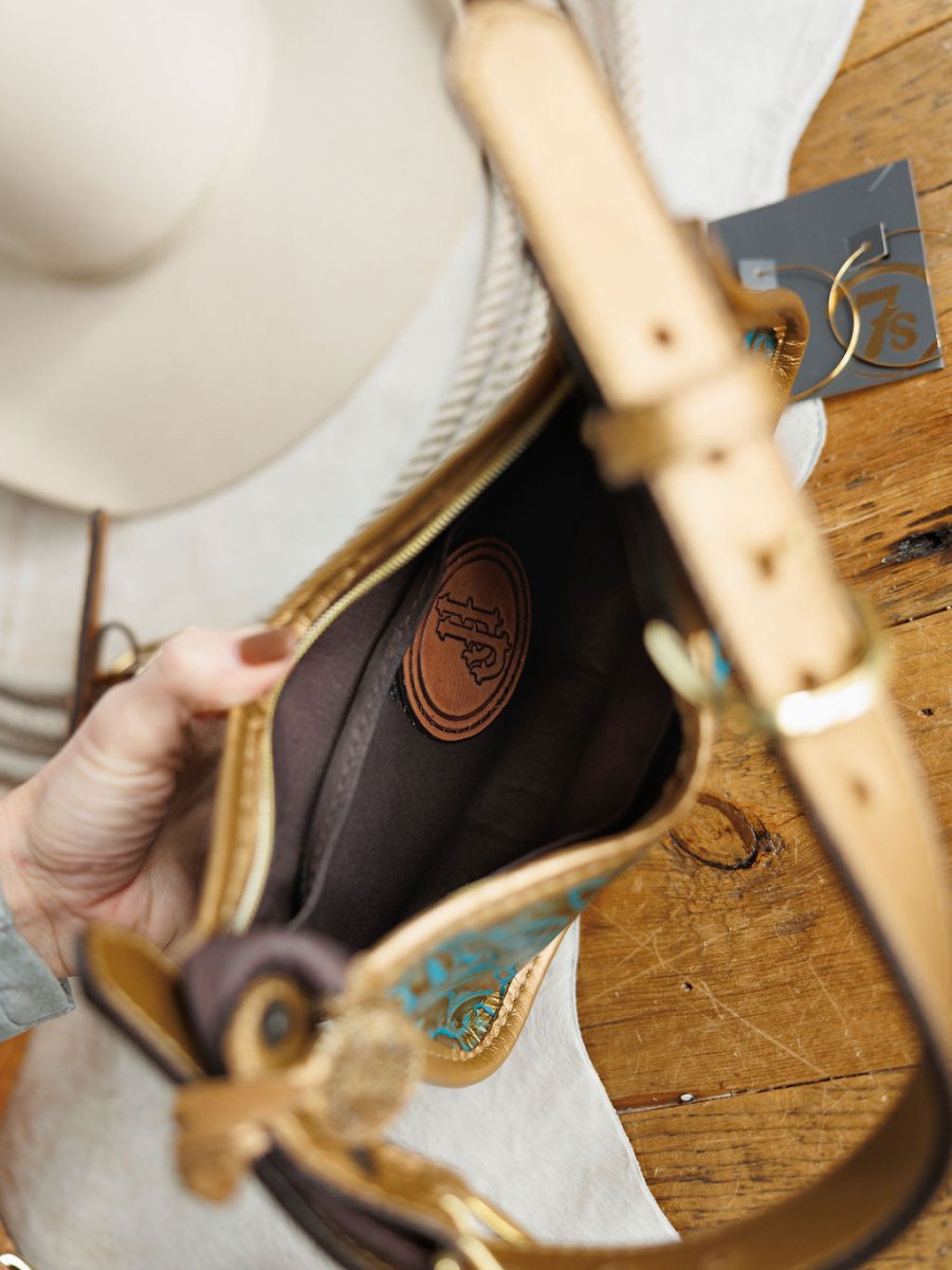 A hand holds open The Ganado, a tan and turquoise tooled leather handbag by Savannah Sevens western life{&}style, showing its brown interior with a stitched logo patch. Nearby, a white hat and gray card sit on the wooden surface.