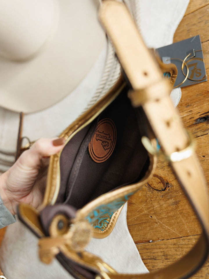 A hand holds open The Ganado, a tan and turquoise tooled leather handbag by Savannah Sevens western life{&}style, showing its brown interior with a stitched logo patch. Nearby, a white hat and gray card sit on the wooden surface.