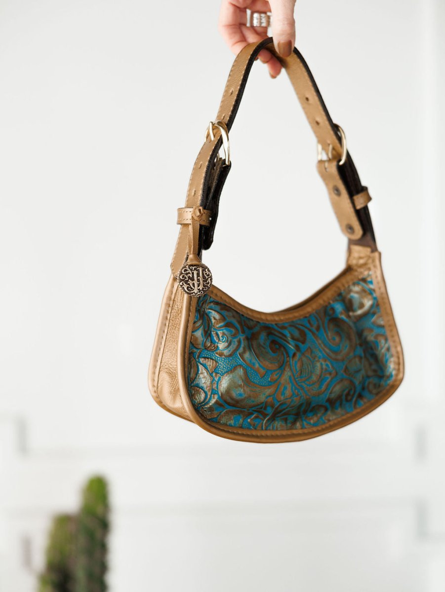 A hand holds The Ganado handbag by Savannah Sevens western life{&}style, featuring tan and turquoise tooled leather with an embossed floral pattern, black accents, and a round metallic charm. A blurred green plant appears against the white background.