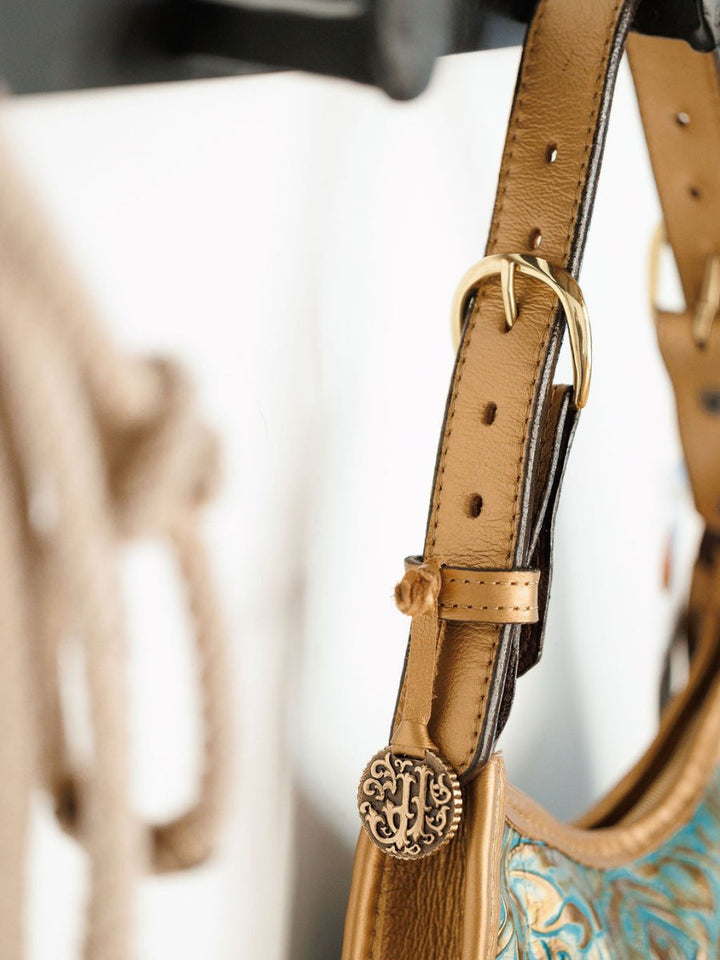 Close-up of The Ganado handbag strap by Savannah Sevens western life{&}style, showcasing hand-tooled leather, a gold buckle, and an ornate round metal charm with intricate design. Blurred background.
