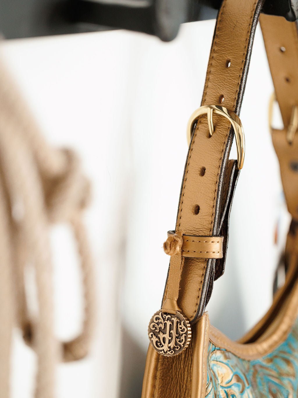 Close-up of The Ganado handbag strap by Savannah Sevens western life{&}style, showcasing hand-tooled leather, a gold buckle, and an ornate round metal charm with intricate design. Blurred background.