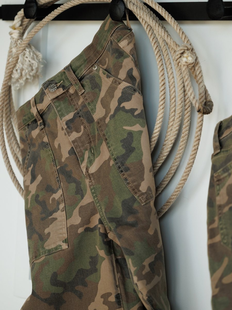 The Granger High Rise Wide Leg Jean by Savannah Sevens western life{&}style features a camo pattern and hangs on a hook, complemented by thick ropes. Its wide-leg cut stands out against the minimalist, neutral-toned backdrop.