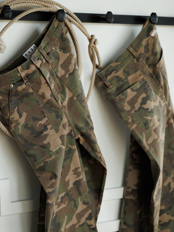 Two pairs of The Granger High Rise Wide Leg Jeans by Savannah Sevens western life{&}style, featuring camo patterns, hang side by side on black hooks against a white wall, with a knotted rope draped among the hooks.