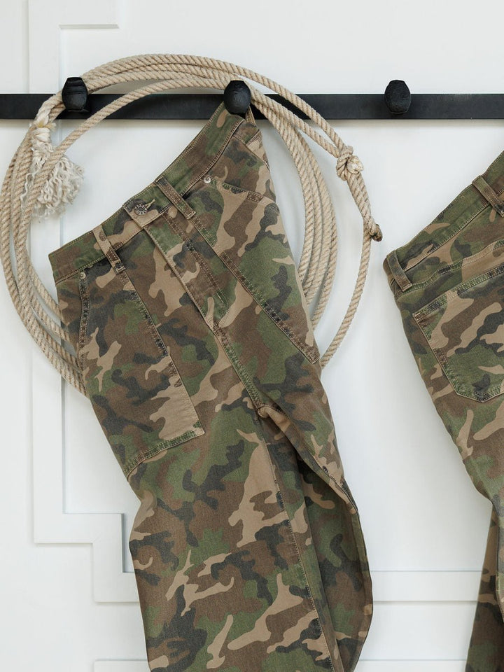 The Granger High Rise Wide Leg Jean by Savannah Sevens western life{&}style, featuring a camo pattern, hangs on a black wall hook with a coiled rope beside it against a white background.