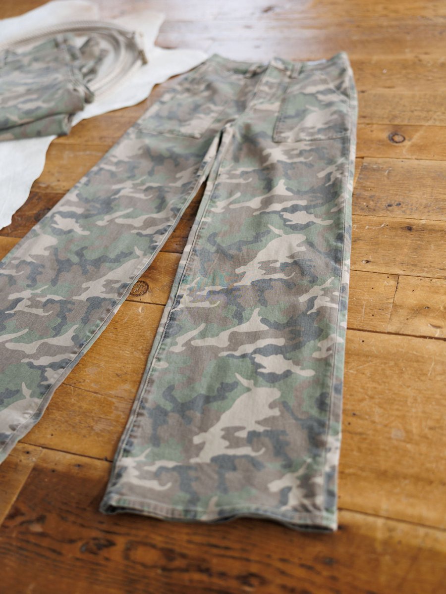 The Granger High Rise Wide Leg Jean by Savannah Sevens western life{&}style in a camo pattern lies flat on a wooden floor beside another folded garment.