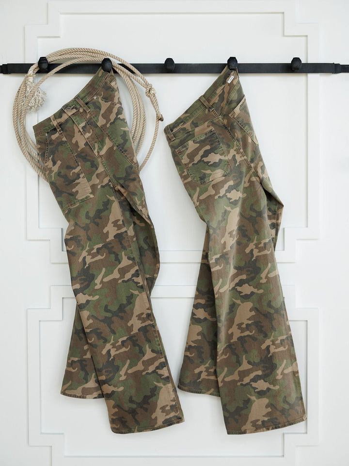 Two pairs of The Granger High Rise Wide Leg Jeans by Savannah Sevens western life{&}style in a camo print hang on black hooks against a white wall, with a coiled rope on the hook to the left of the jeans.