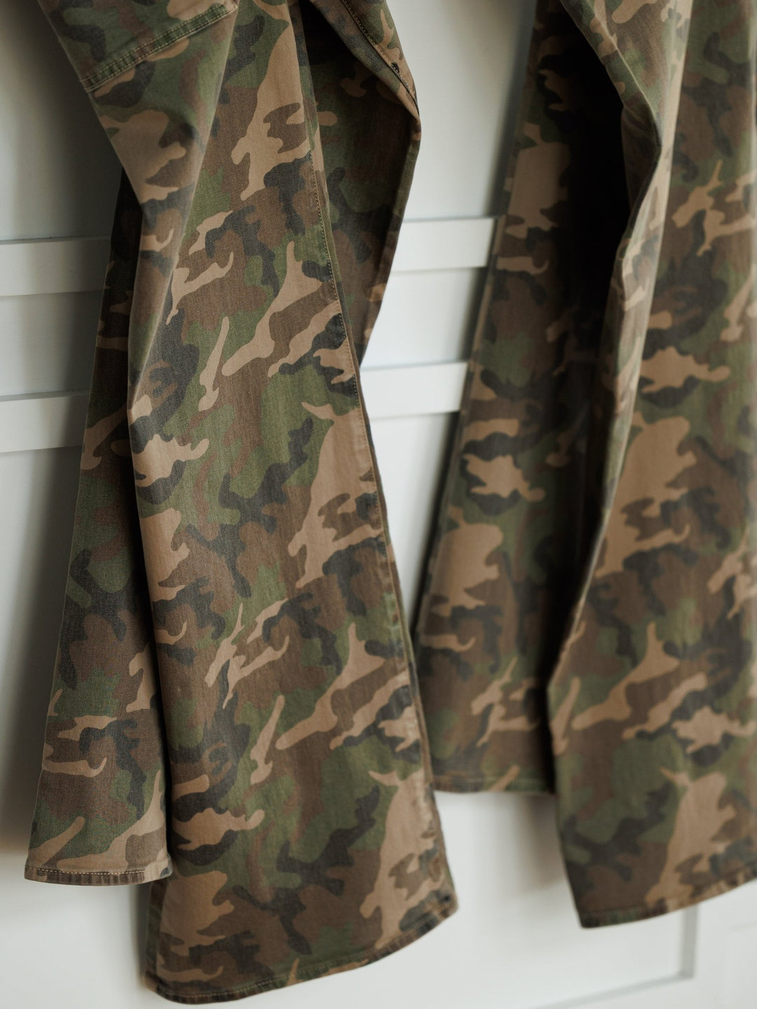 Two pairs of The Granger High Rise Wide Leg Jean by Savannah Sevens western life{&}style, in camo pattern denim, hang side by side on a white wall or door.