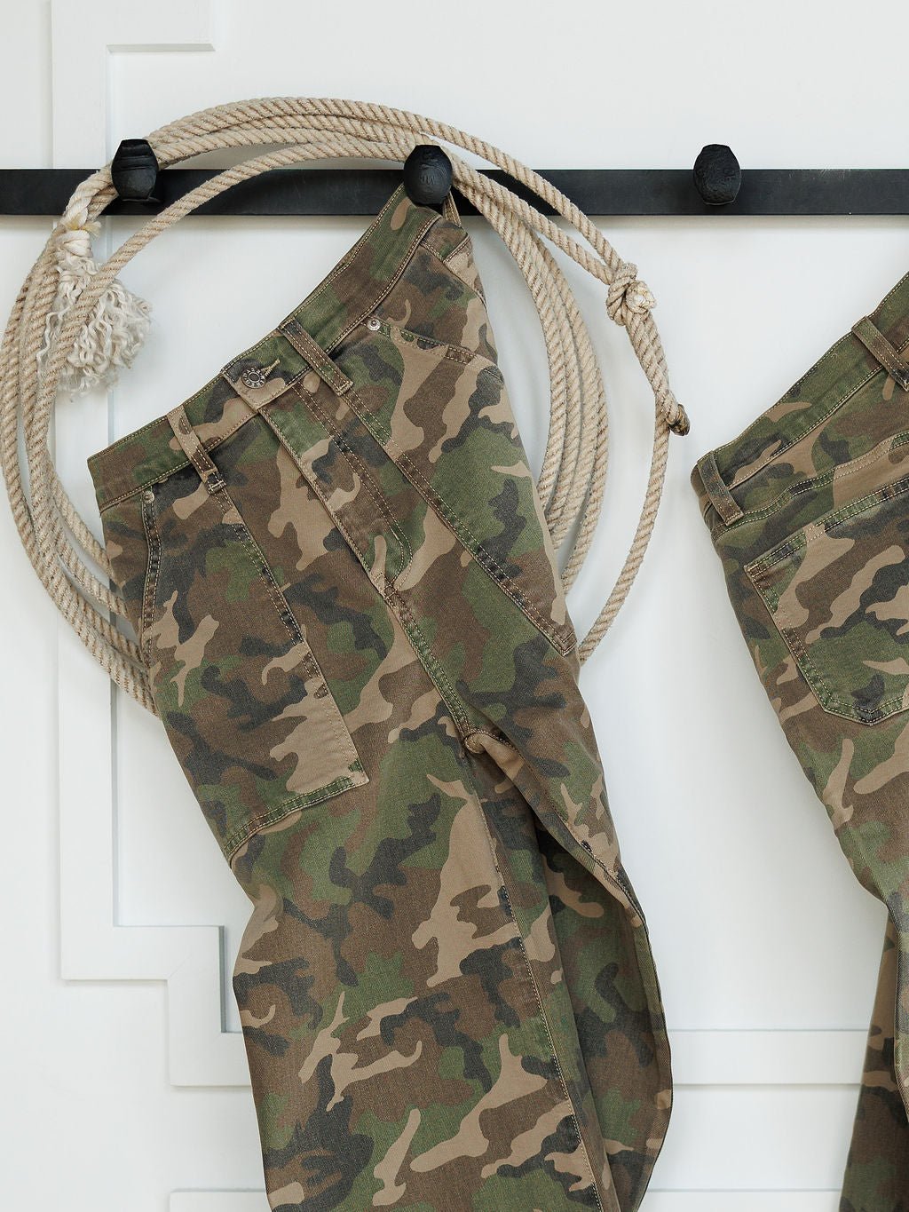 The Granger High Rise Wide Leg Jean by Savannah Sevens western life{&}style, featuring a camo pattern, hangs on a black wall hook with a coiled rope beside it against a white background.