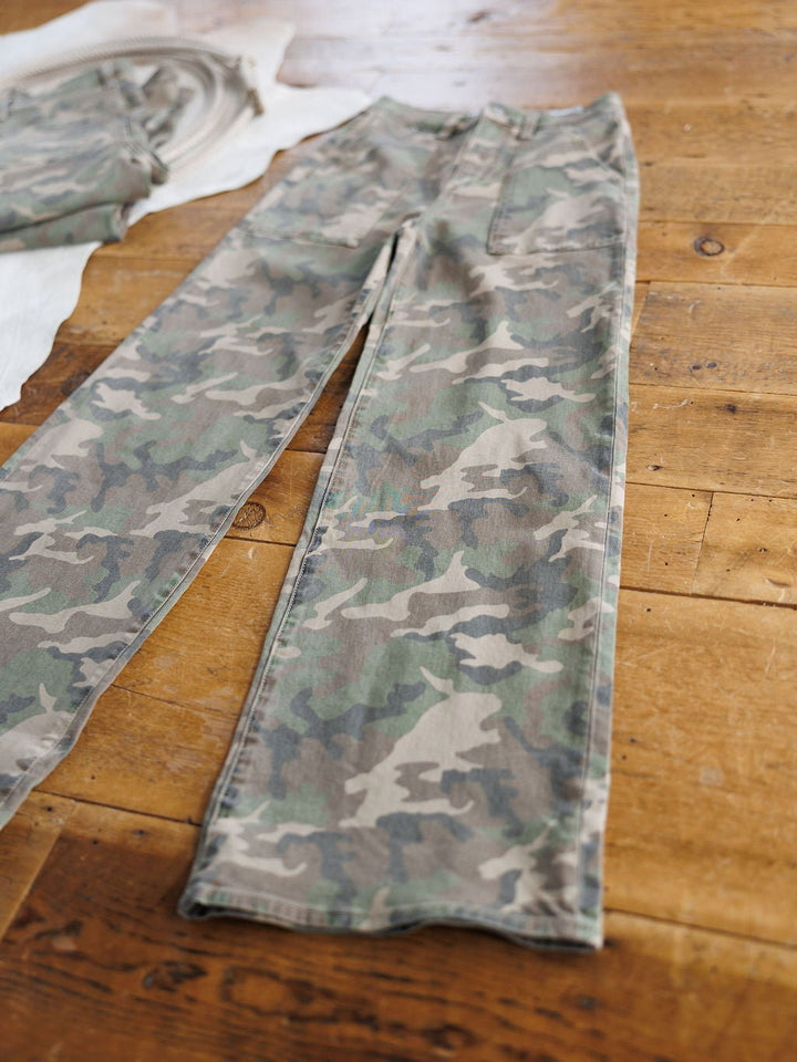 The Granger High Rise Wide Leg Jean by Savannah Sevens western life{&}style in a camo pattern lies flat on a wooden floor beside another folded garment.