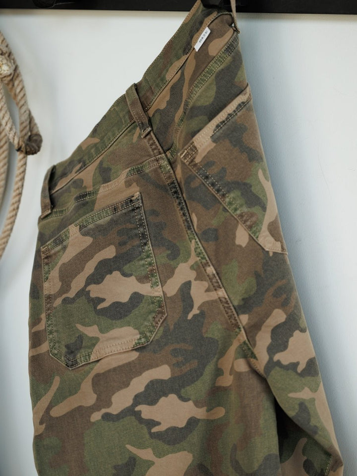 The Granger High Rise Wide Leg Jean by Savannah Sevens western life{&}style in a camo pattern hangs on a black rack near a coiled rope, set against a light-colored wall.