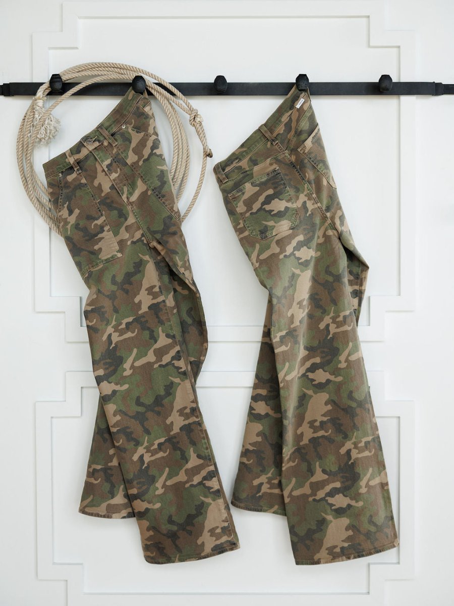 Two pairs of The Granger High Rise Wide Leg Jeans by Savannah Sevens western life{&}style in a camo print hang on black hooks against a white wall, with a coiled rope on the hook to the left of the jeans.