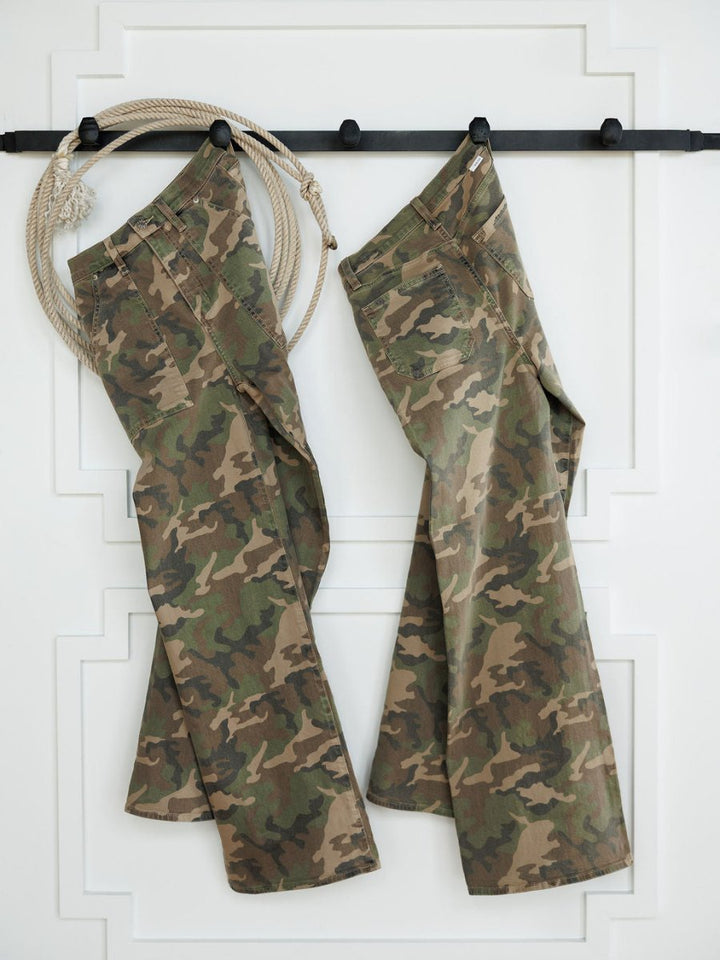 Two pairs of The Granger High Rise Wide Leg Jeans by Savannah Sevens western life{&}style in a camo print hang on black hooks against a white wall, with a coiled rope on the hook to the left of the jeans.