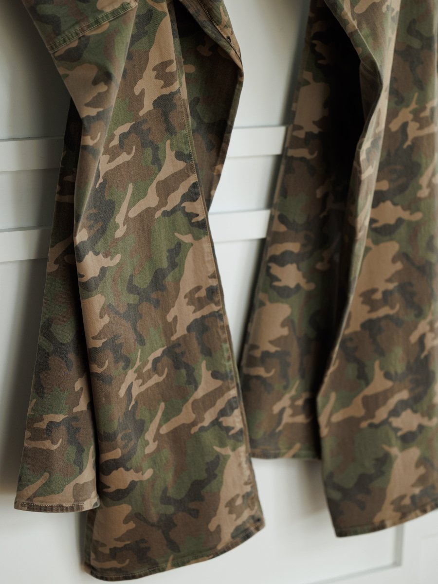 Two pairs of The Granger High Rise Wide Leg Jean by Savannah Sevens western life{&}style, in camo pattern denim, hang side by side on a white wall or door.