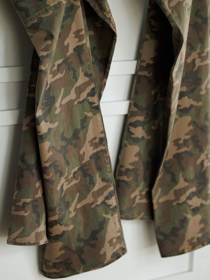 Two pairs of The Granger High Rise Wide Leg Jean by Savannah Sevens western life{&}style, in camo pattern denim, hang side by side on a white wall or door.