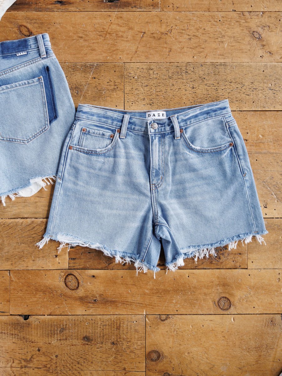 The Hayden Shorts by Savannah Sevens western life{&}style, light blue denim with a raw cut hem, are displayed flat on a wooden floor next to the edge of another similar pair of jeans.