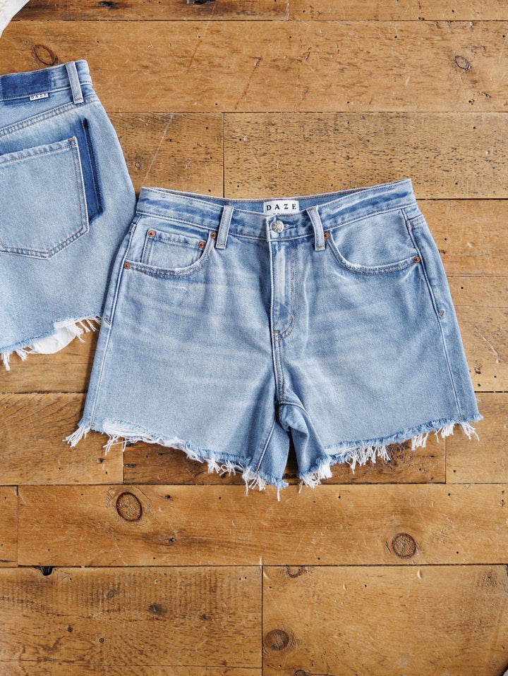 The Hayden Shorts by Savannah Sevens western life{&}style, light blue denim with a raw cut hem, are displayed flat on a wooden floor next to the edge of another similar pair of jeans.