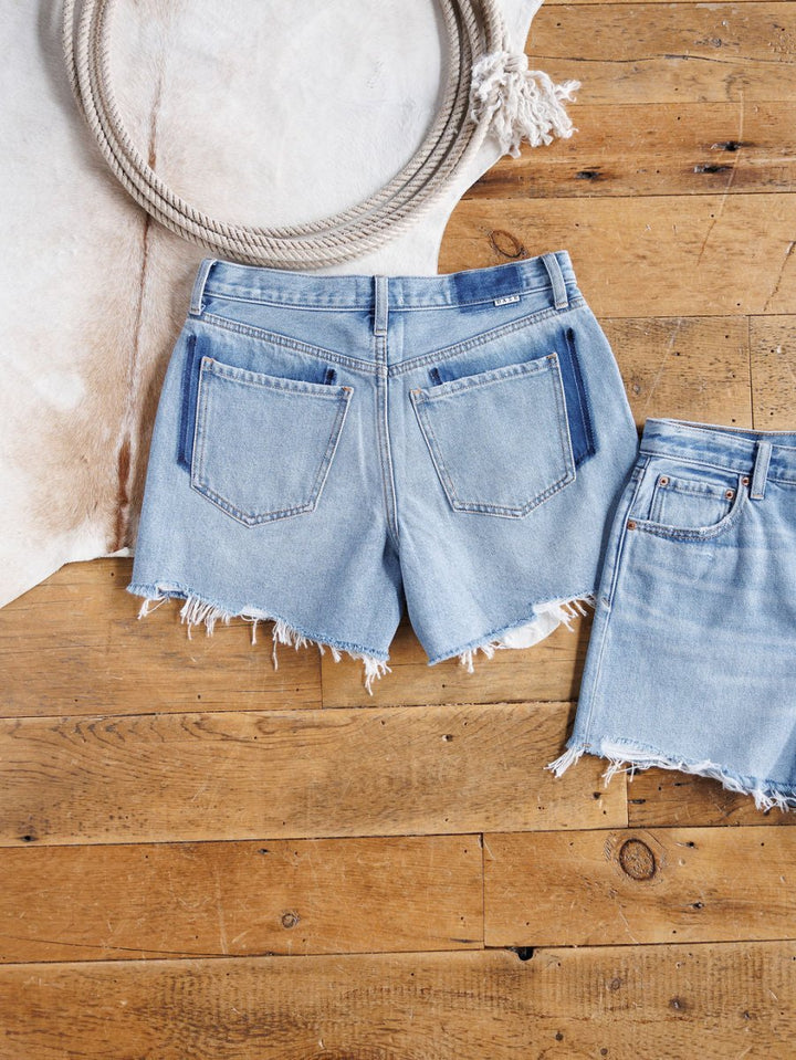 Two pairs of The Hayden Shorts by Savannah Sevens western life{&}style, featuring a relaxed fit, light blue denim, frayed hems, and visible back pockets, are displayed on a wooden floor beside a coiled rope and cowhide rug.
