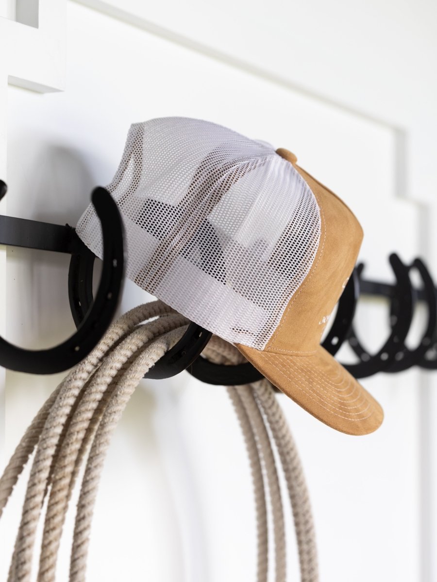 A tan and white Savannah Sevens western life{&}style The Hell I Won't Cap and a coiled rope hang on black horseshoe-shaped hooks mounted on a white wall.