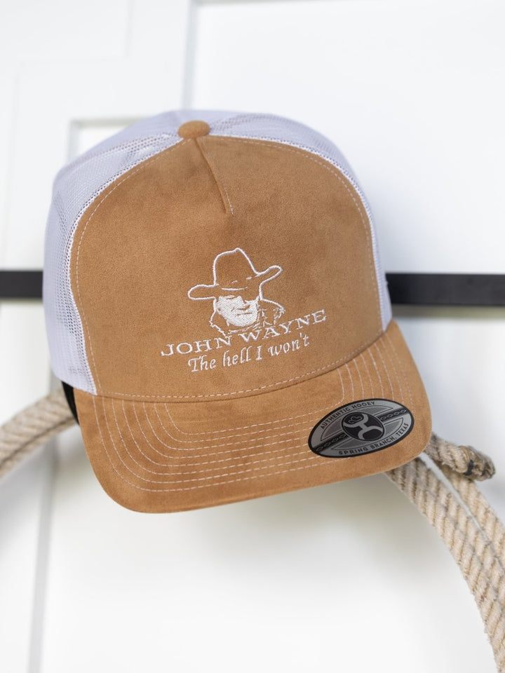 The Hell I Won't Cap by Savannah Sevens western life{&}style features a brown and white design, embroidered cowboy face, "JOHN WAYNE The hell I won't" text, and a bill sticker. It hangs on a white surface above a coiled rope, adding Western charm.