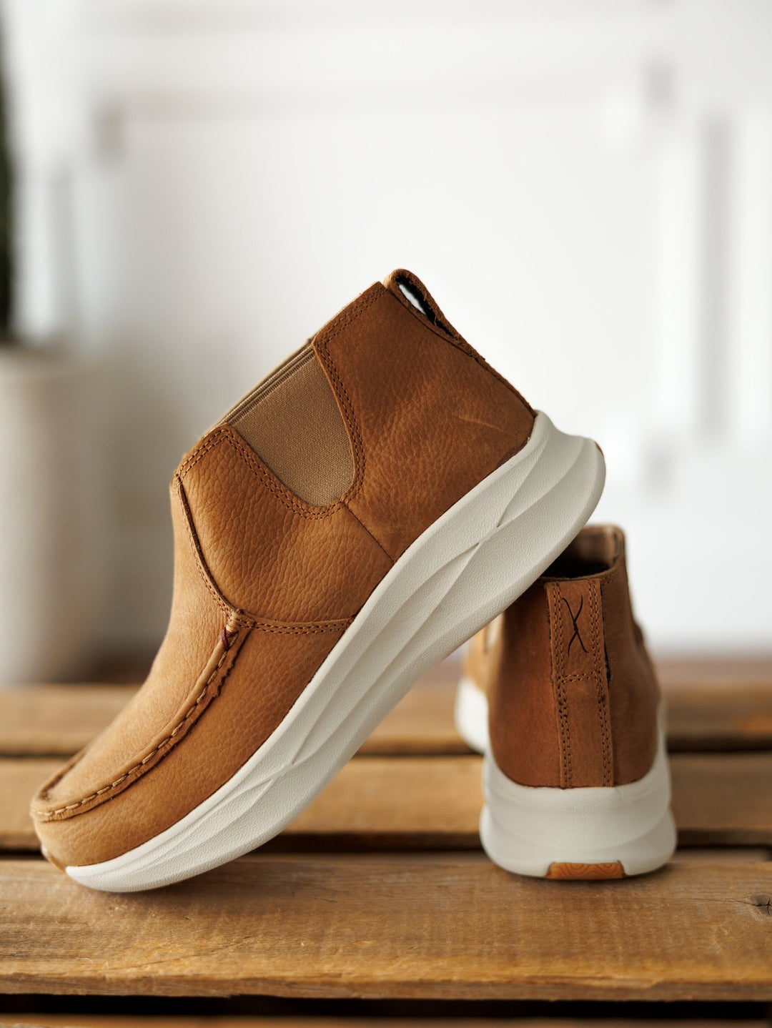 A pair of Savannah Sevens western life{&}style The Holden brown leather casual slip-ons with white slip-resistant soles are shown on a wooden surface—one upright, the other tilted to display the side and sole against a softly blurred background. By TwistedX.