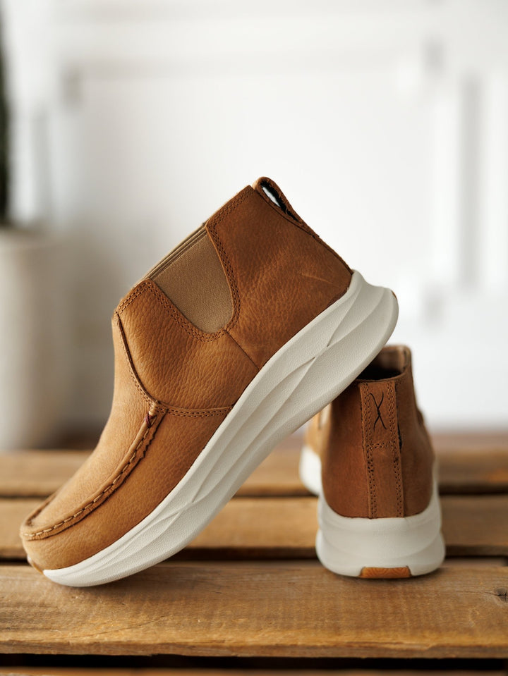 A pair of Savannah Sevens western life{&}style The Holden brown leather casual slip-ons with white slip-resistant soles are shown on a wooden surface—one upright, the other tilted to display the side and sole against a softly blurred background. By TwistedX.