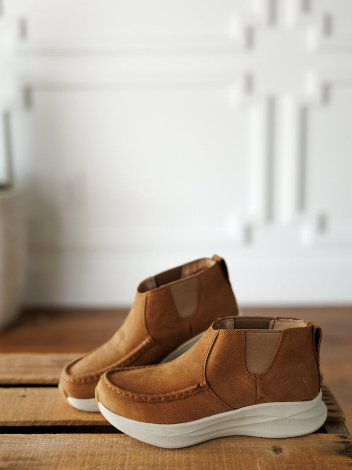 A pair of The Holden tan leather casual shoes by Savannah Sevens western life{&}style with thick white soles are placed on a wooden surface. The background is a white, textured wall with geometric patterns. Made by Twisted X. 