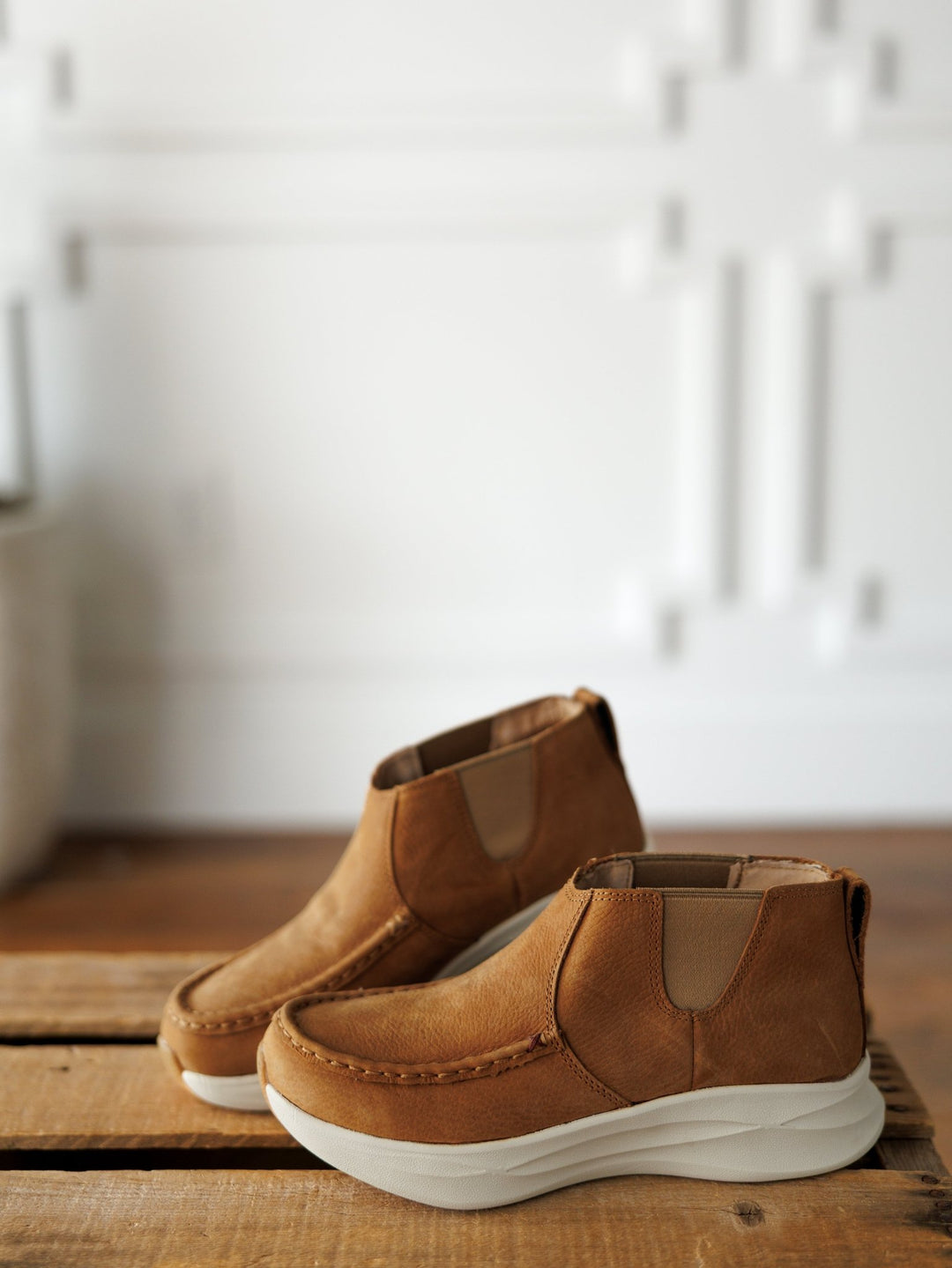 A pair of The Holden tan leather casual shoes by Savannah Sevens western life{&}style with thick white soles are placed on a wooden surface. The background is a white, textured wall with geometric patterns. Made by Twisted X. 