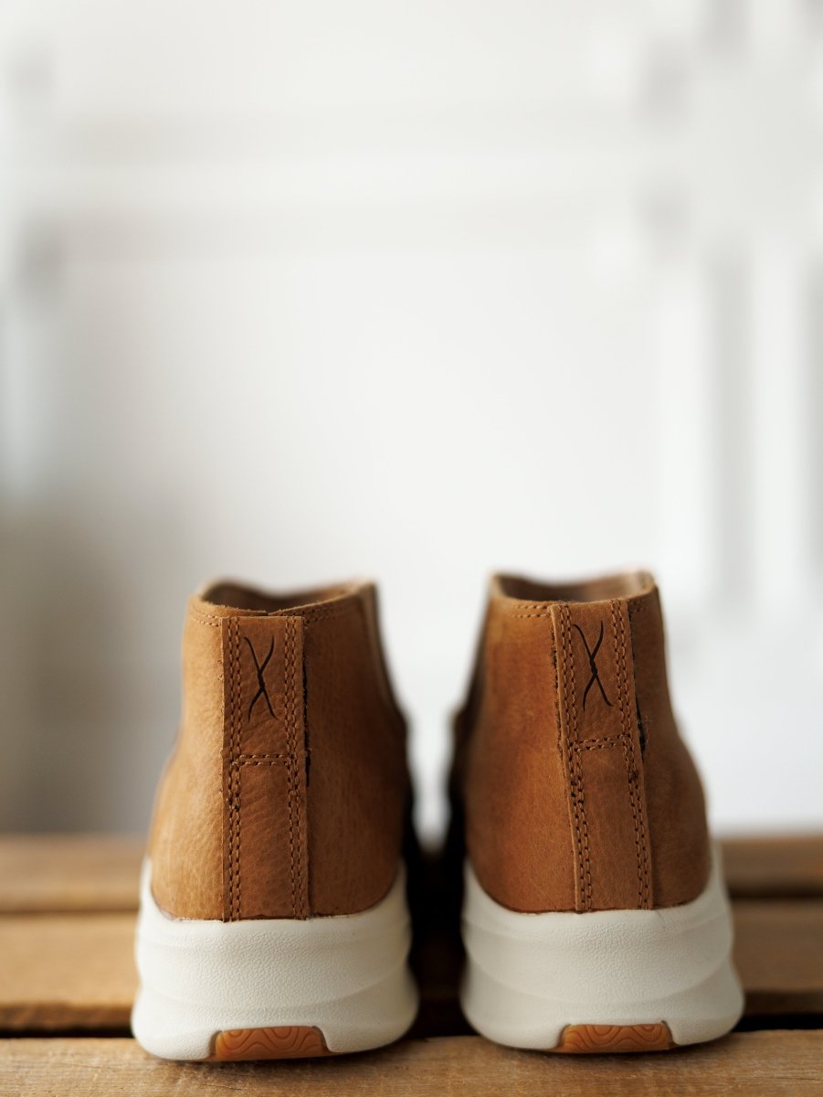 The Holden by Savannah Sevens western life{&}style: light brown suede sneakers with white rubber soles, shown from the back on wood with a blurred white background. Slip-resistant for comfort and casual style.