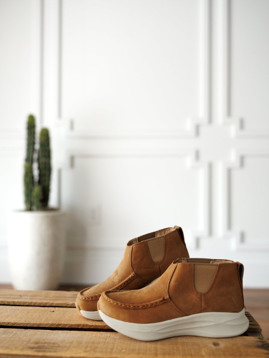 A pair of Savannah Sevens western life{&}style The Holden brown leather casual shoes with thick white, slip-resistant soles sit on a wooden surface, with a potted cactus and a white paneled wall in the background. By Twisted X.