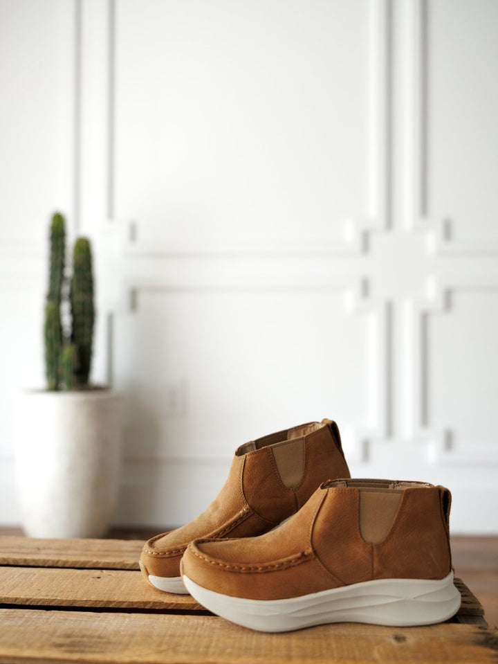 A pair of Savannah Sevens western life{&}style The Holden brown leather casual shoes with thick white, slip-resistant soles sit on a wooden surface, with a potted cactus and a white paneled wall in the background. By Twisted X.