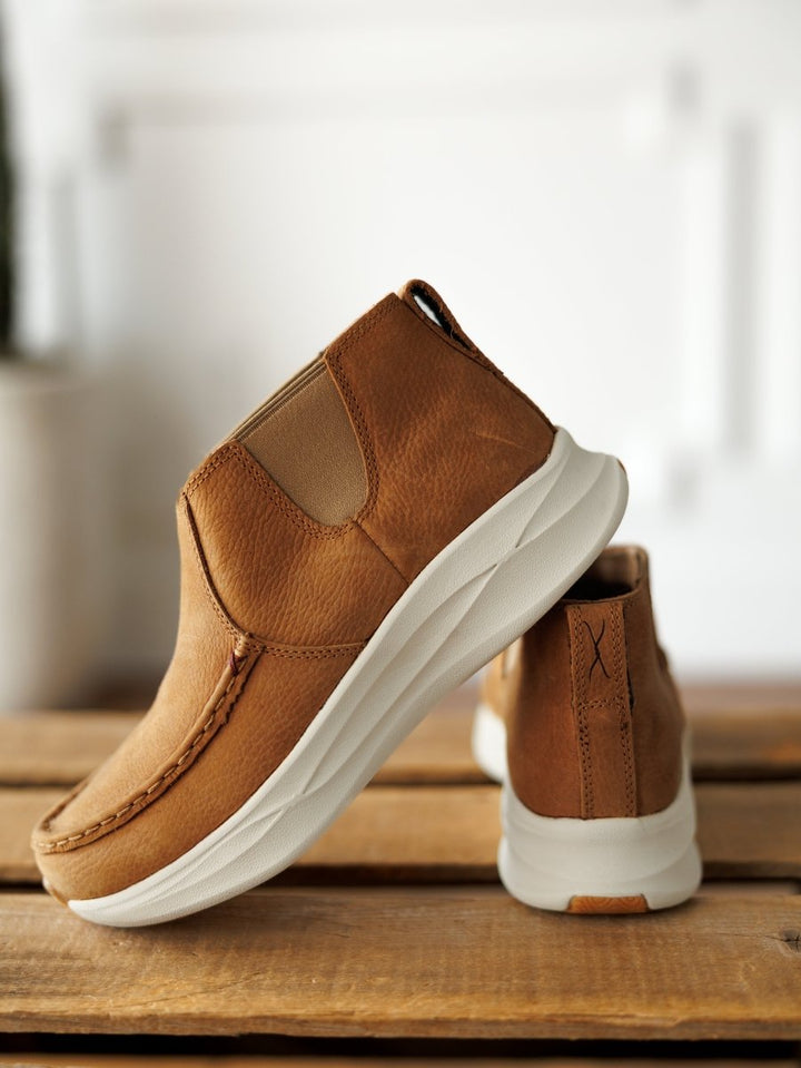 A pair of Savannah Sevens western life{&}style The Holden brown leather casual slip-ons with white slip-resistant soles are shown on a wooden surface—one upright, the other tilted to display the side and sole against a softly blurred background. By TwistedX.