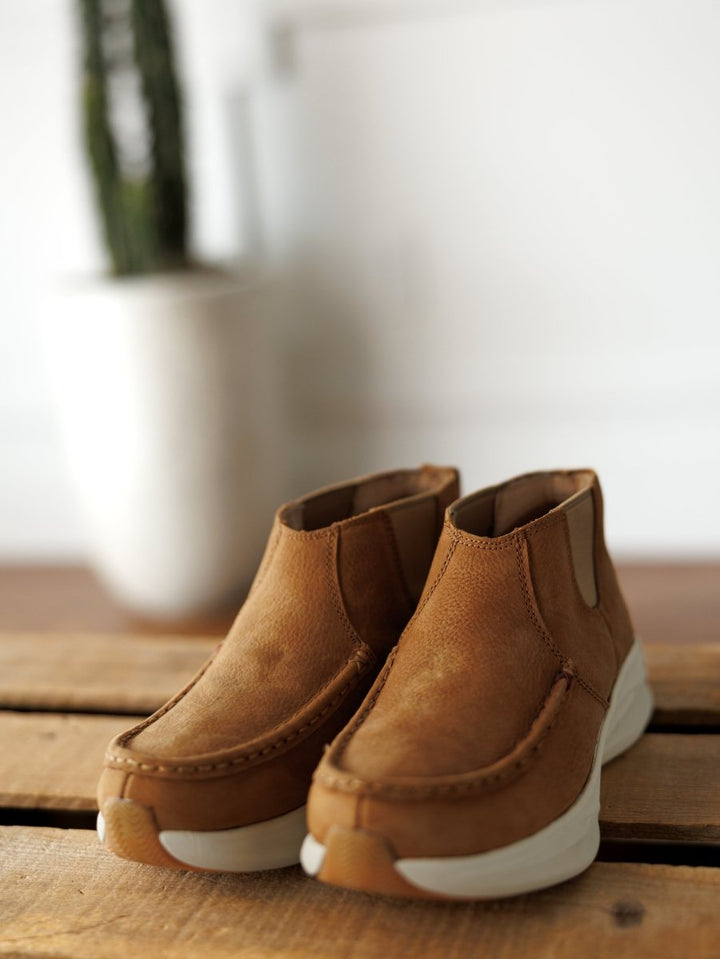 A pair of The Holden tan leather casual shoes by Savannah Sevens western life{&}style with white soles sits on a wooden surface; in the blurred background, a white planter holds a green cactus. By Twisted X.