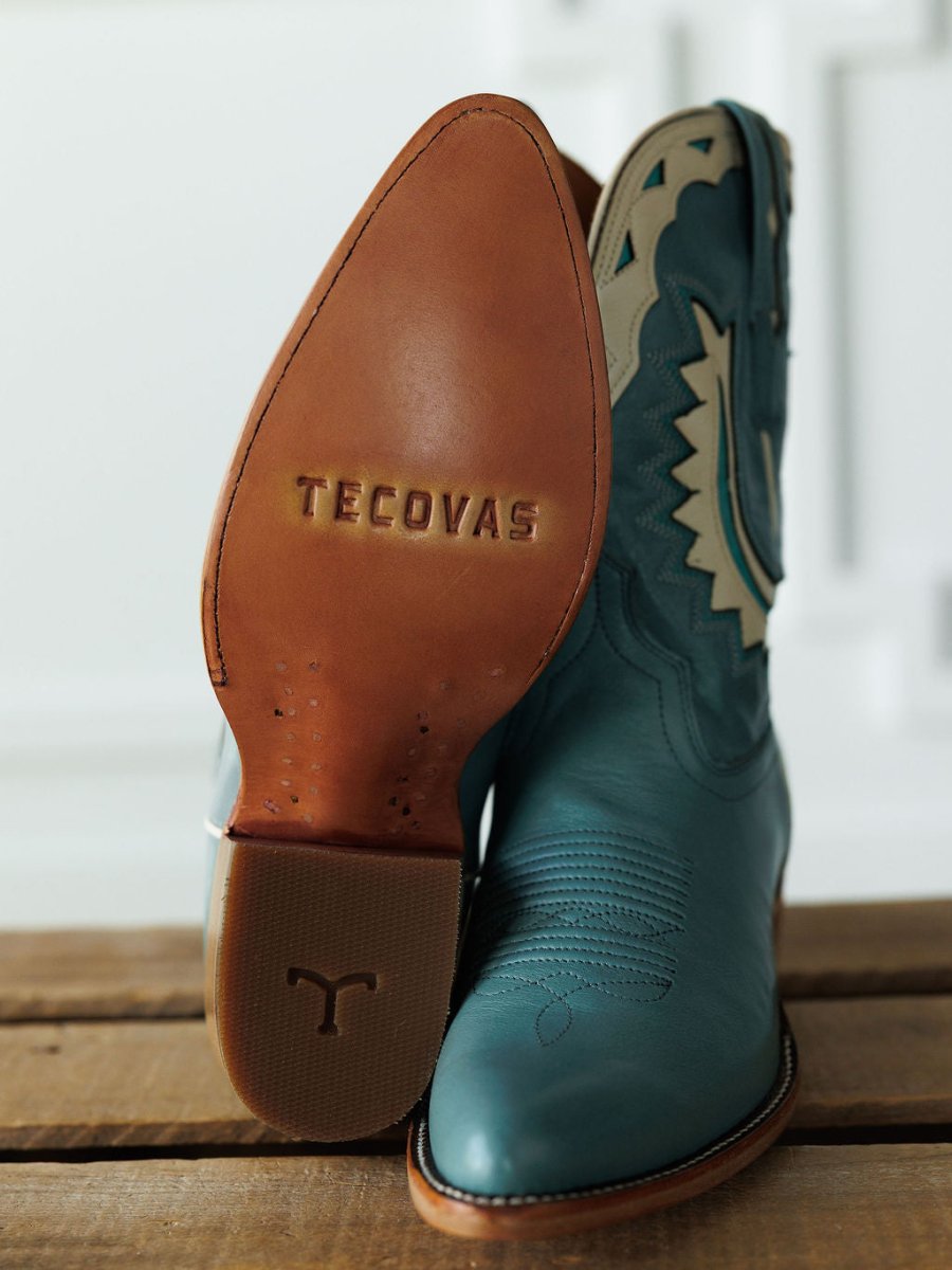 A close-up of The Jolene Boot {Seafoam} by Savannah Sevens western life{&}style, one upright and the other showing the tan sole with a Y-shaped logo, both set on a wooden surface. Boots handmade by Tecovas.