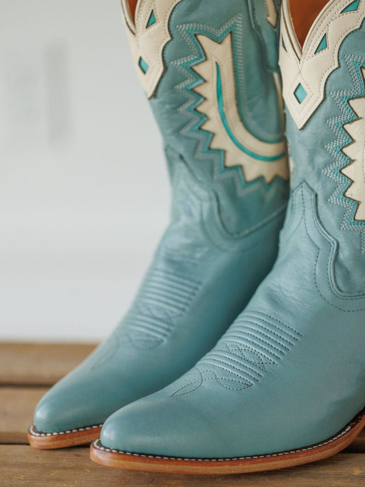 A close-up of The Jolene Boot {Seafoam} by Savannah Sevens western life{&}style in turquoise leather with cream and teal stitching and patterns, resting on a wooden surface. Boots handmade by Tecovas.