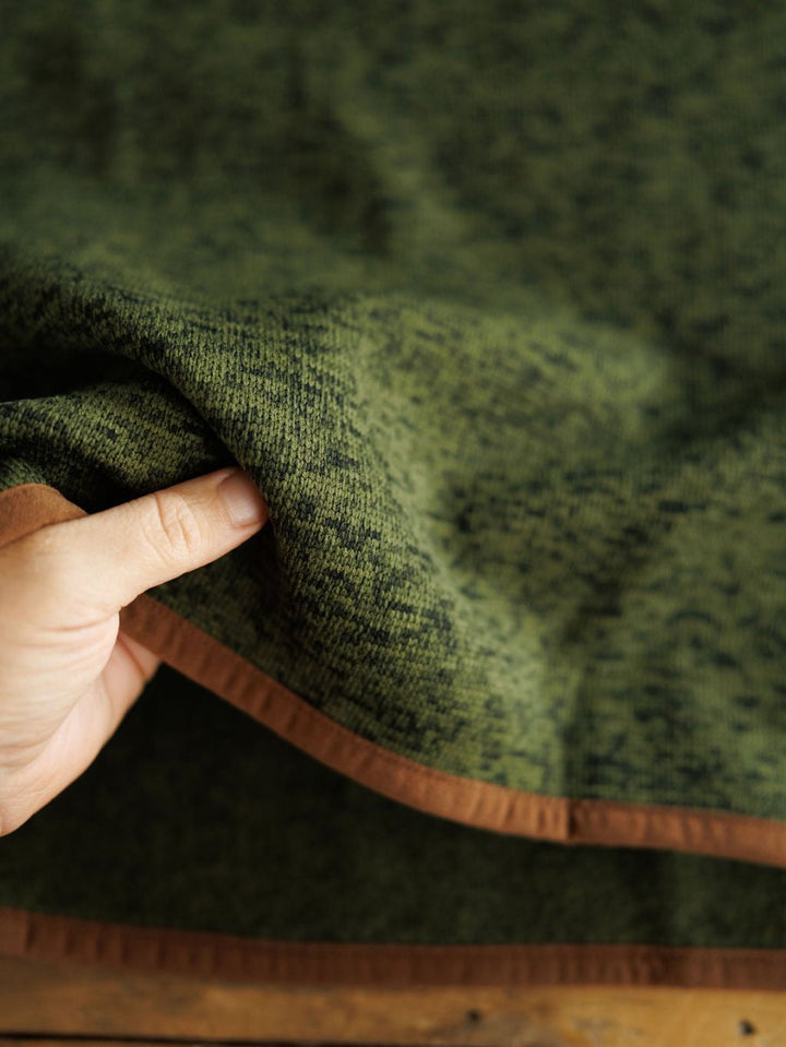 A hand lifts the edge of The Kempton {Men's} by Savannah Sevens western life{&}style, a green, fleece-lined blanket with brown trim, showing off its softness and thickness.