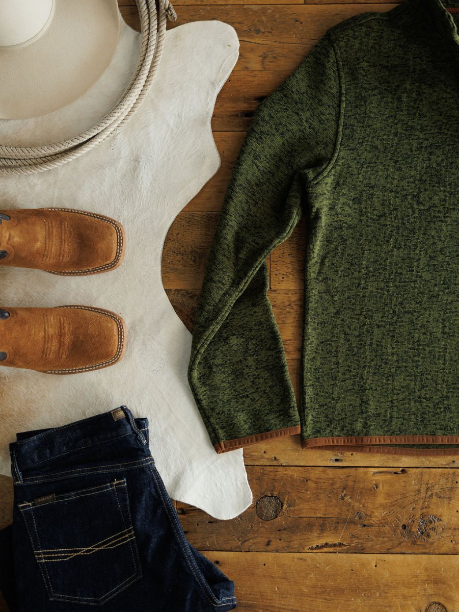 The Kempton {Men's} sweater from Savannah Sevens western life{&}style is styled with dark blue jeans, brown cowboy boots, a cowboy hat, and a coiled rope on a wooden surface with a white cowhide rug.