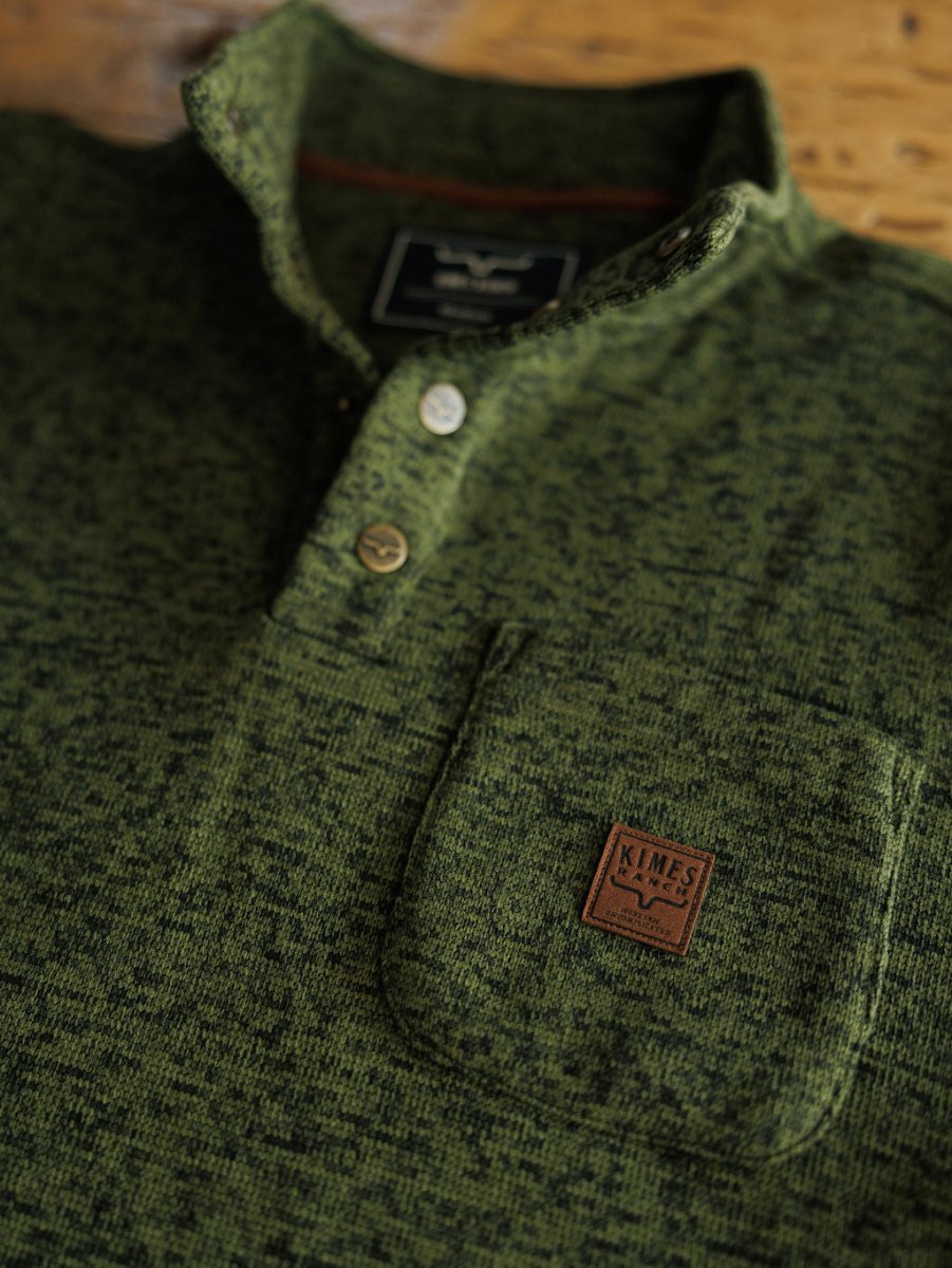Close-up of The Kempton men's henley by Savannah Sevens western life{&}style, featuring green textured fabric, buttons, a chest pocket with a brown leather patch embossed with the Texas emblem. The pullover is laid flat on a wooden surface.