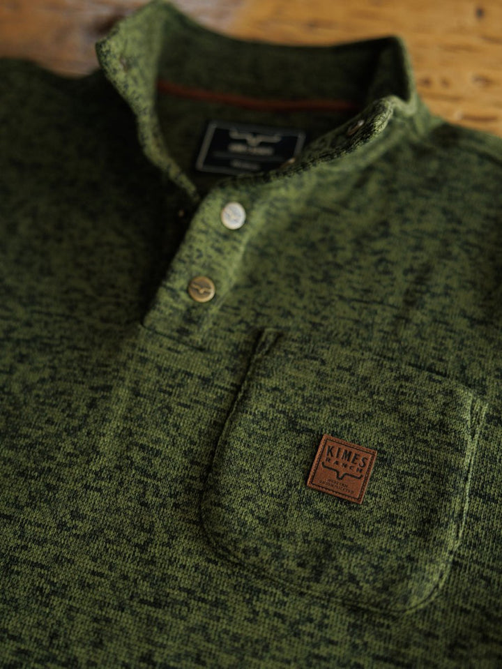 Close-up of The Kempton men's henley by Savannah Sevens western life{&}style, featuring green textured fabric, buttons, a chest pocket with a brown leather patch embossed with the Texas emblem. The pullover is laid flat on a wooden surface.
