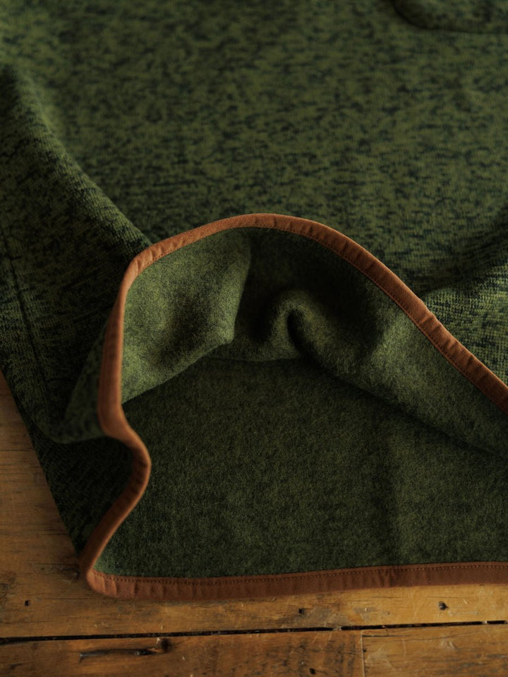 A close-up of a green wool blanket with brown trim, folded at the corner on a wooden surface—ideal for pairing with your favorite Savannah Sevens western life{&}style The Kempton {Men's} pullover.
