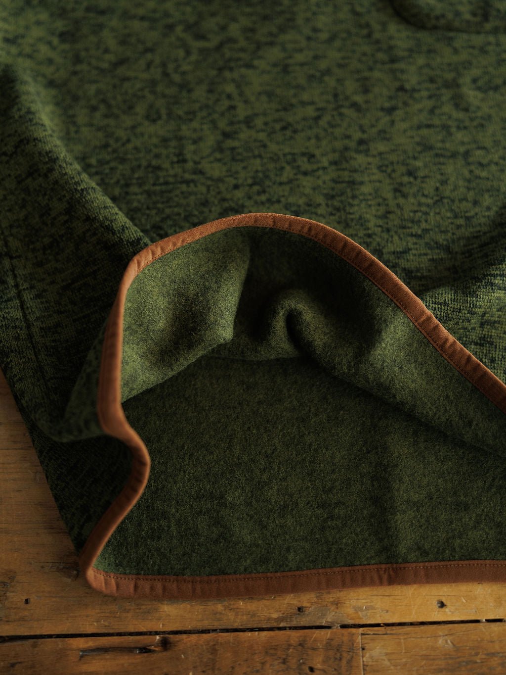 A close-up of a green wool blanket with brown trim, folded at the corner on a wooden surface—ideal for pairing with your favorite Savannah Sevens western life{&}style The Kempton {Men's} pullover.