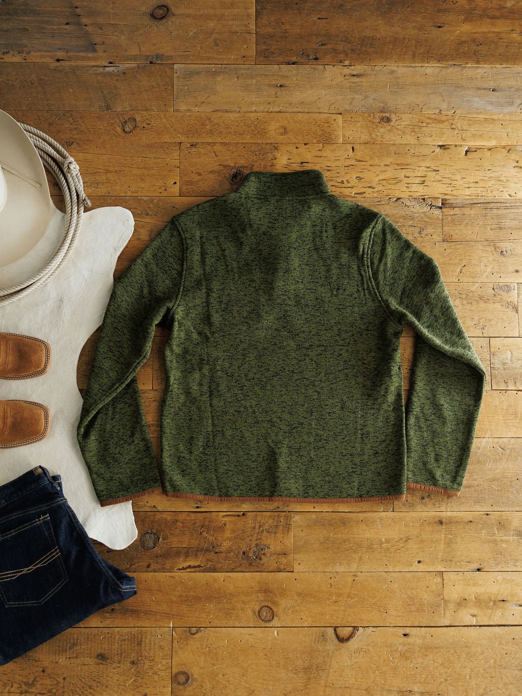 The Kempton {Men's} pullover by Savannah Sevens western life{&}style is displayed flat on wood with jeans, boots, a white cowboy hat, rope, and a cowhide rug for a rugged western look.