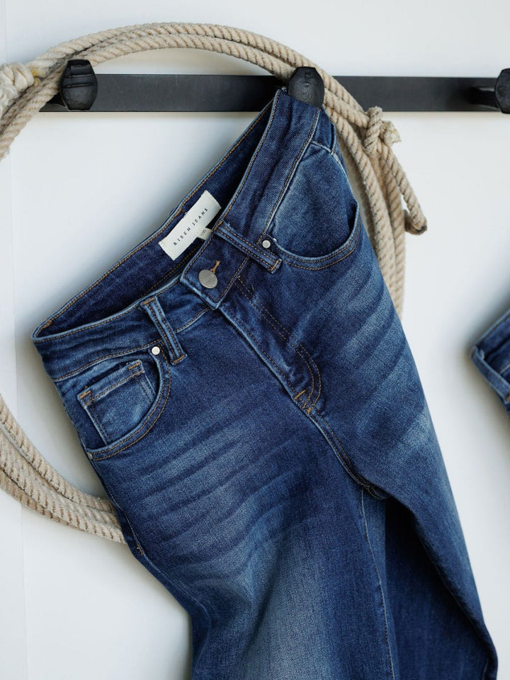 The Knox High Rise Wide Leg Jean by Savannah Sevens western life{&}style, in a medium dark wash with faded detailing and waistband label, hangs from a thick rope on a black wall hook against a white background.