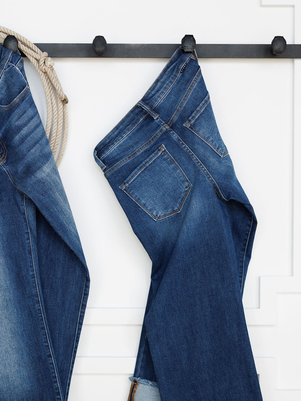 A pair of medium dark wash blue jeans hangs by a belt loop on a black hook against a white wall. On the left, Savannah Sevens western life{&}style's The Knox High Rise Wide Leg Jean and a coiled rope are partially visible.