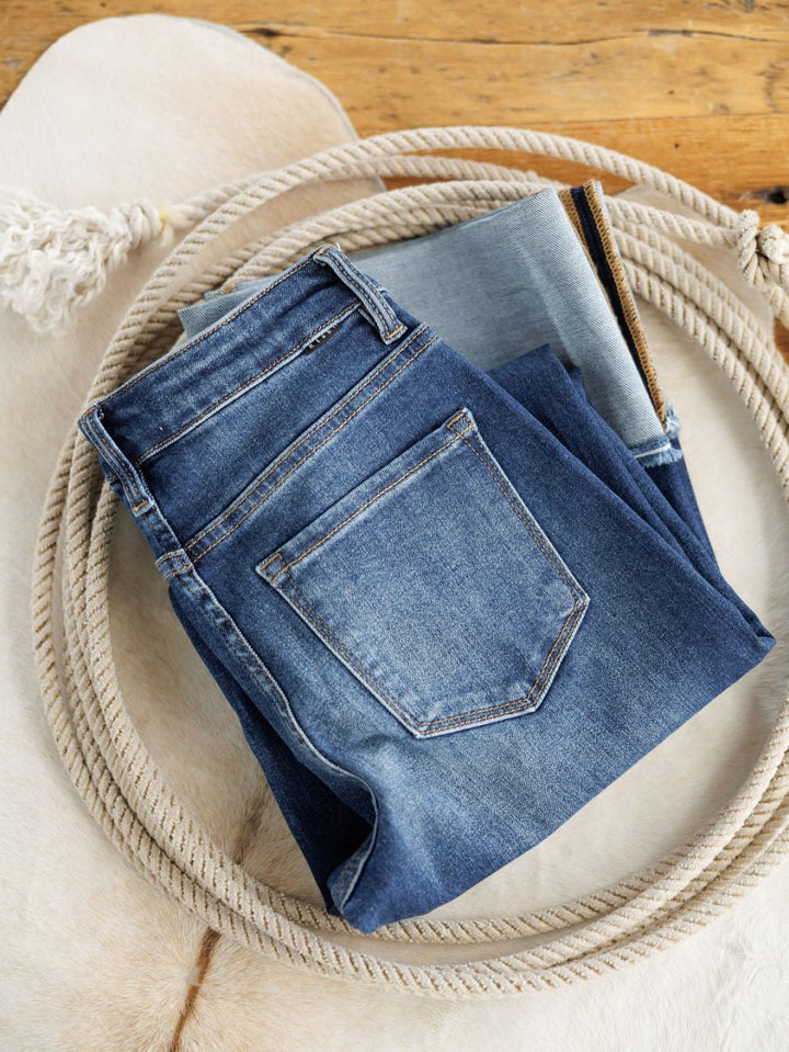 The Knox High Rise Wide Leg Jean from Savannah Sevens western life{&}style in a medium dark wash is displayed inside a coiled rope on a white fur rug, with a wooden floor in the background.