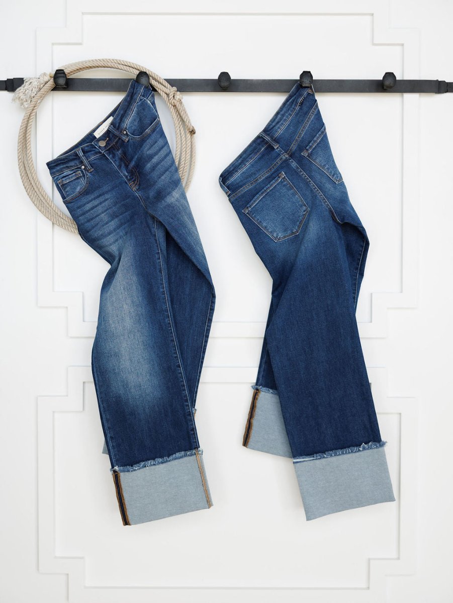 Two pairs of The Knox High Rise Wide Leg Jean by Savannah Sevens western life{&}style, in medium dark wash with cuffed hems, hang side by side on a black rod against a white wall. A thick rope is looped around the rod next to the jeans.