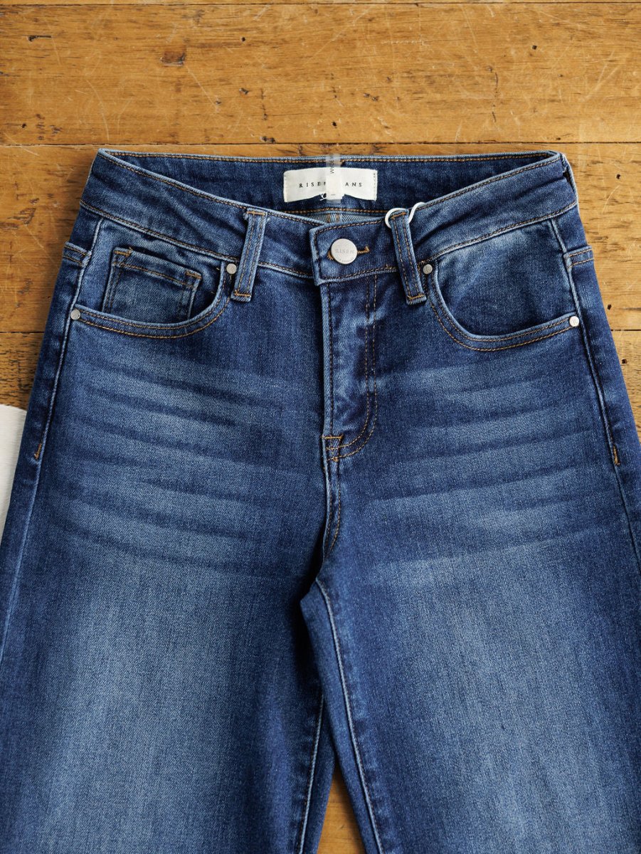 The Knox High Rise Wide Leg Jean by Savannah Sevens western life{&}style in medium dark wash lies flat on wood, showing the waistband, front pockets, button, zipper, and faded lines across the upper thighs.