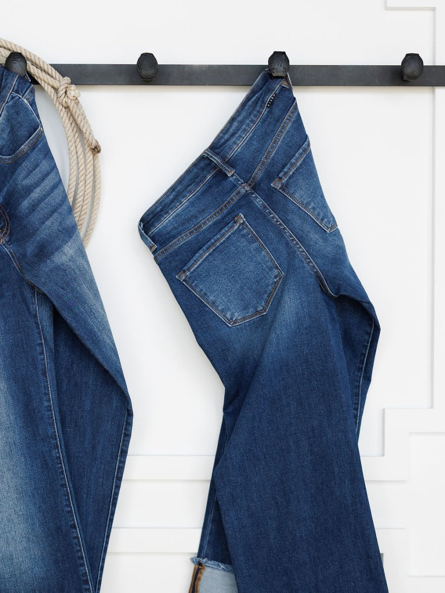 A pair of medium dark wash blue jeans hangs by a belt loop on a black hook against a white wall. On the left, Savannah Sevens western life{&}style's The Knox High Rise Wide Leg Jean and a coiled rope are partially visible.