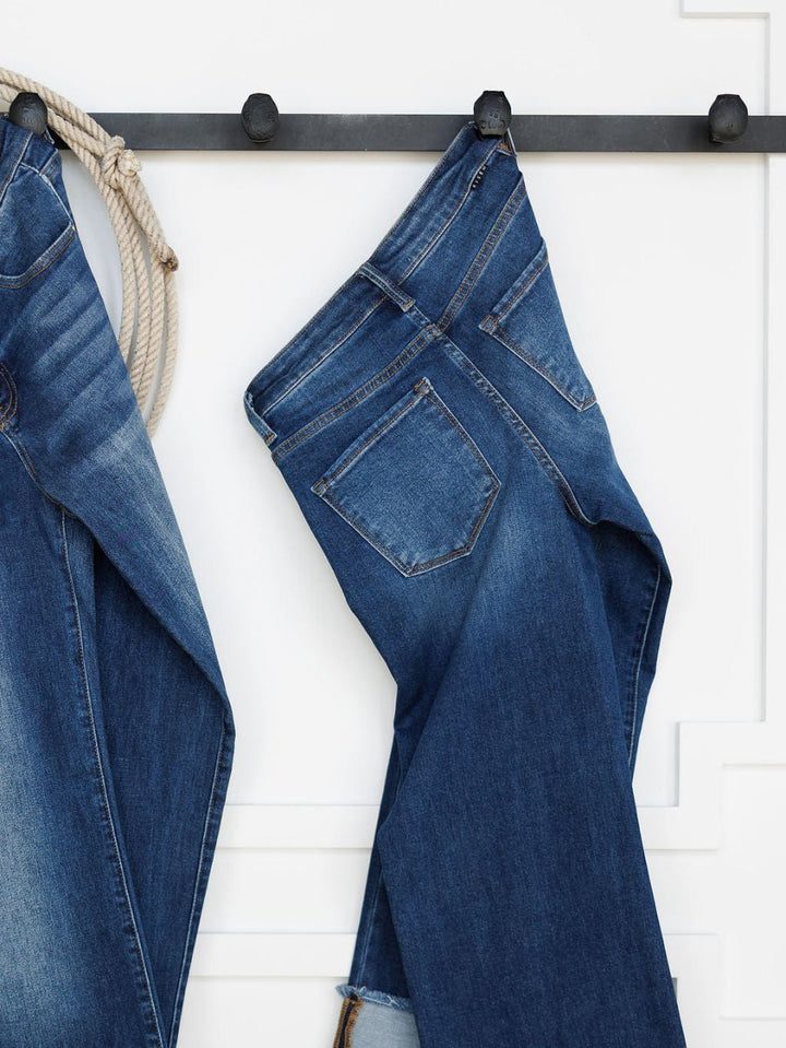 A pair of medium dark wash blue jeans hangs by a belt loop on a black hook against a white wall. On the left, Savannah Sevens western life{&}style's The Knox High Rise Wide Leg Jean and a coiled rope are partially visible.