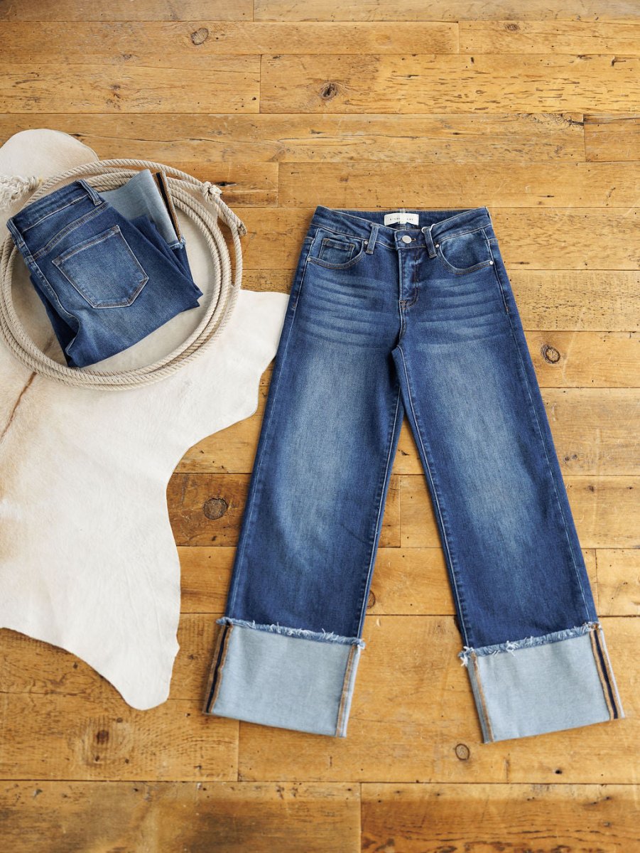 Two pairs of blue jeans from Savannah Sevens western life{&}style on a wooden floor: one folded on a white cowhide rug, the other is The Knox High Rise Wide Leg Jean in medium dark wash with cuffed legs and frayed hems.