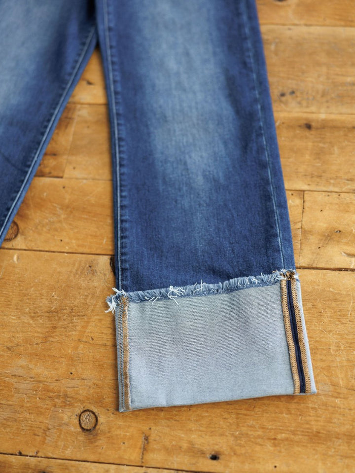 The Knox High Rise Wide Leg Jean by Savannah Sevens western life{&}style in a medium dark wash features frayed cuffs and folded legs that reveal the lighter inside fabric, displayed on a wooden surface.